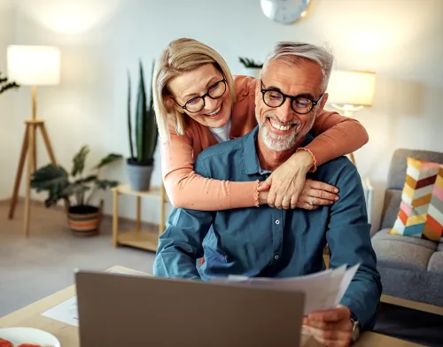 Retired couple hugging and laughing while browsing on their laptop